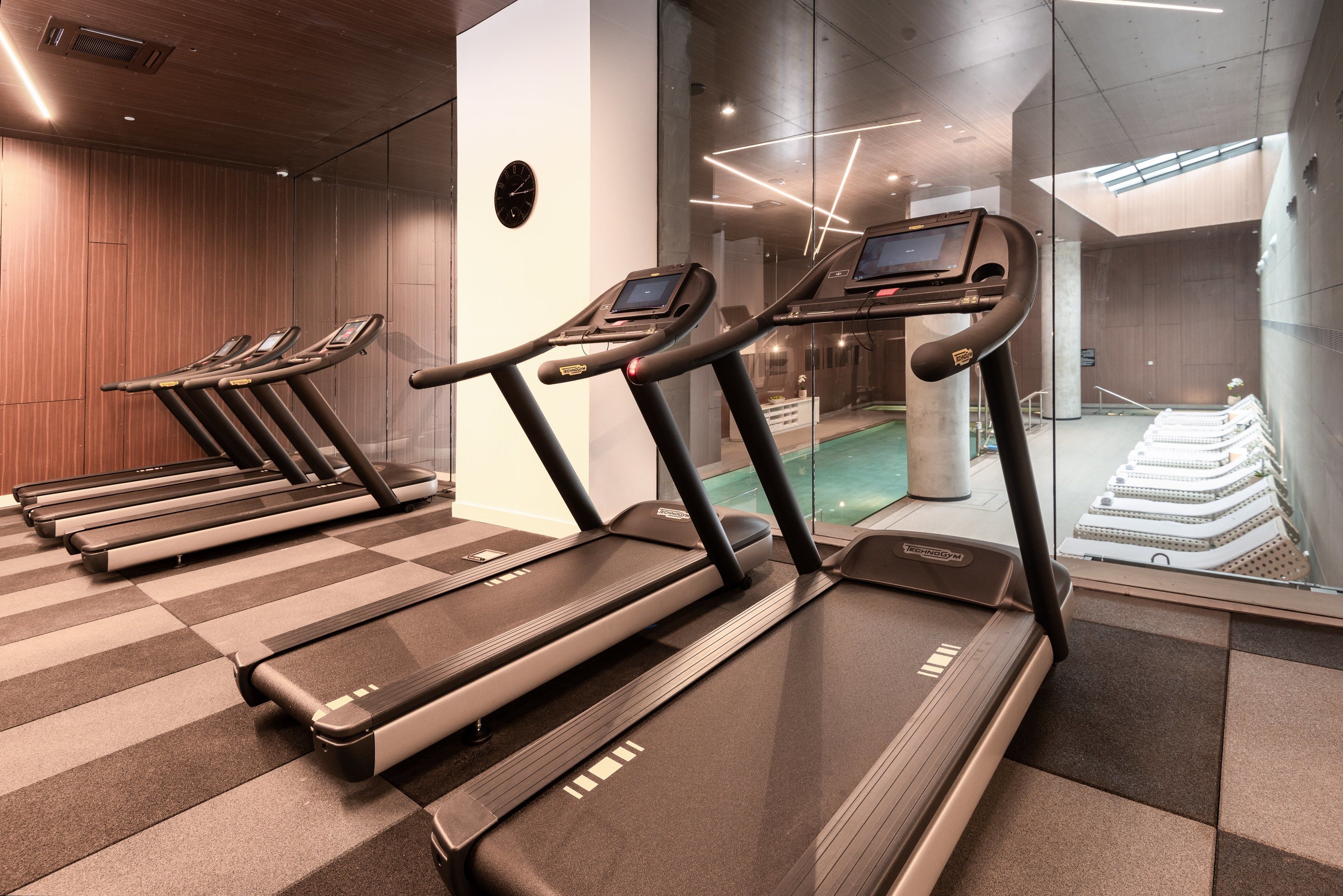 Un gimnasio con numerosas cintas de correr, una zona de piscina a la vista y un diseño arquitectónico moderno.