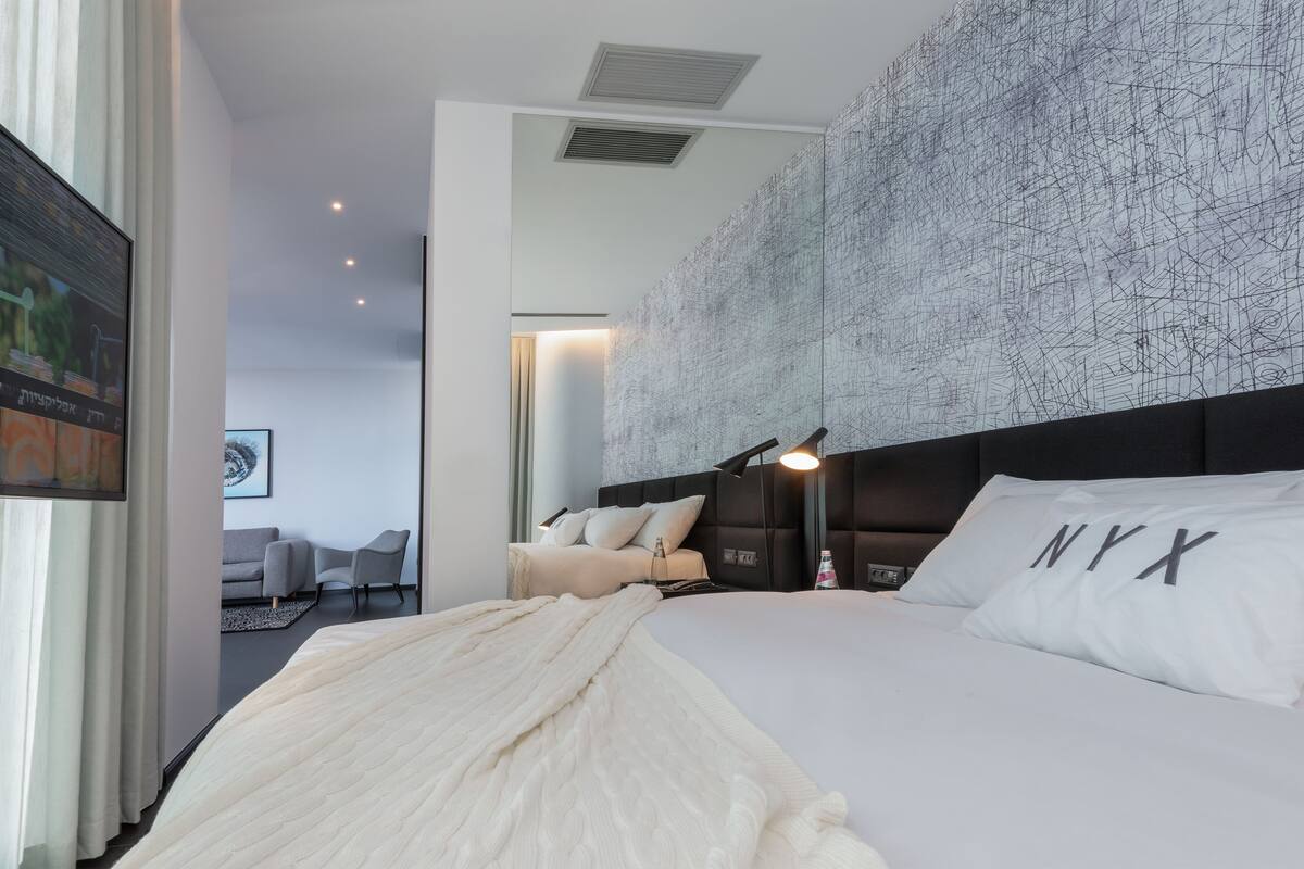 Ein modernes Hotelzimmer mit einem großen Bett, einer Bettablage mit 'NYX'-Kissen, einer Wand mit abstrakter Kunst und Blick auf den angrenzenden Wohnbereich.