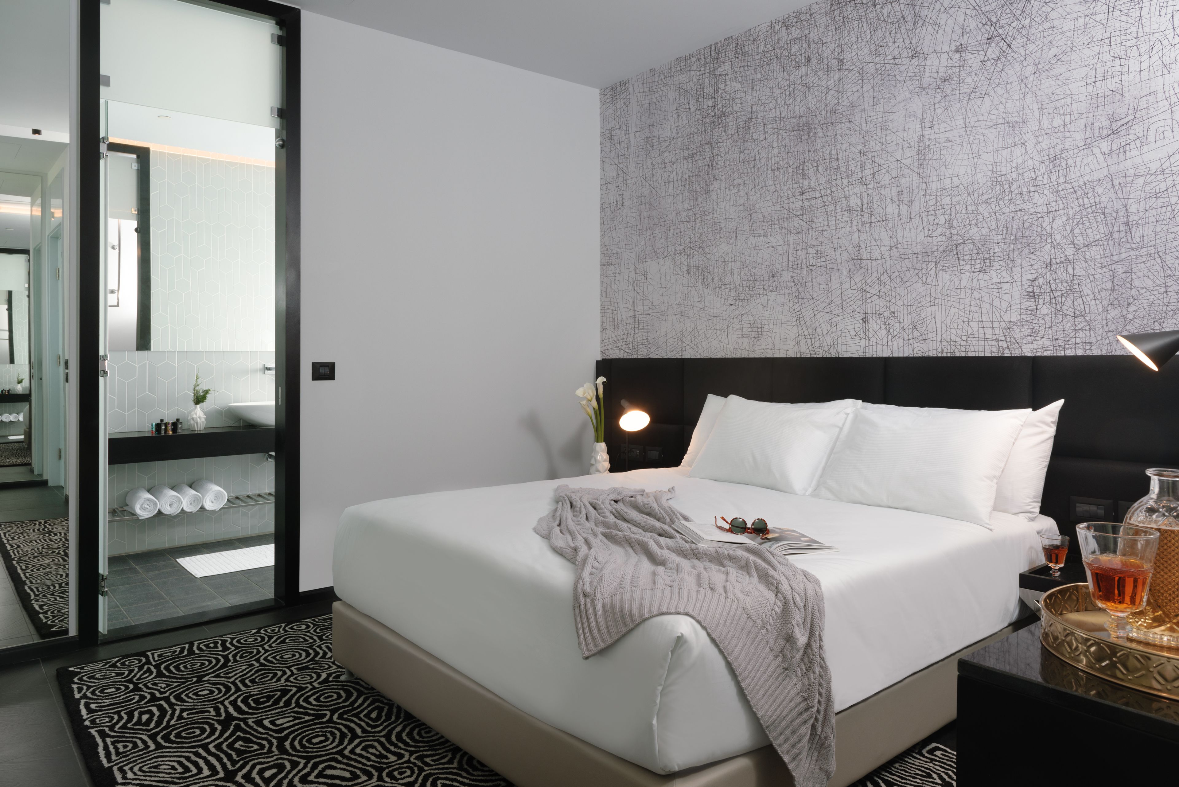 A modern hotel room with a large bed, a nightstand, a glass of beverage, and a bathroom visible through a sliding door.