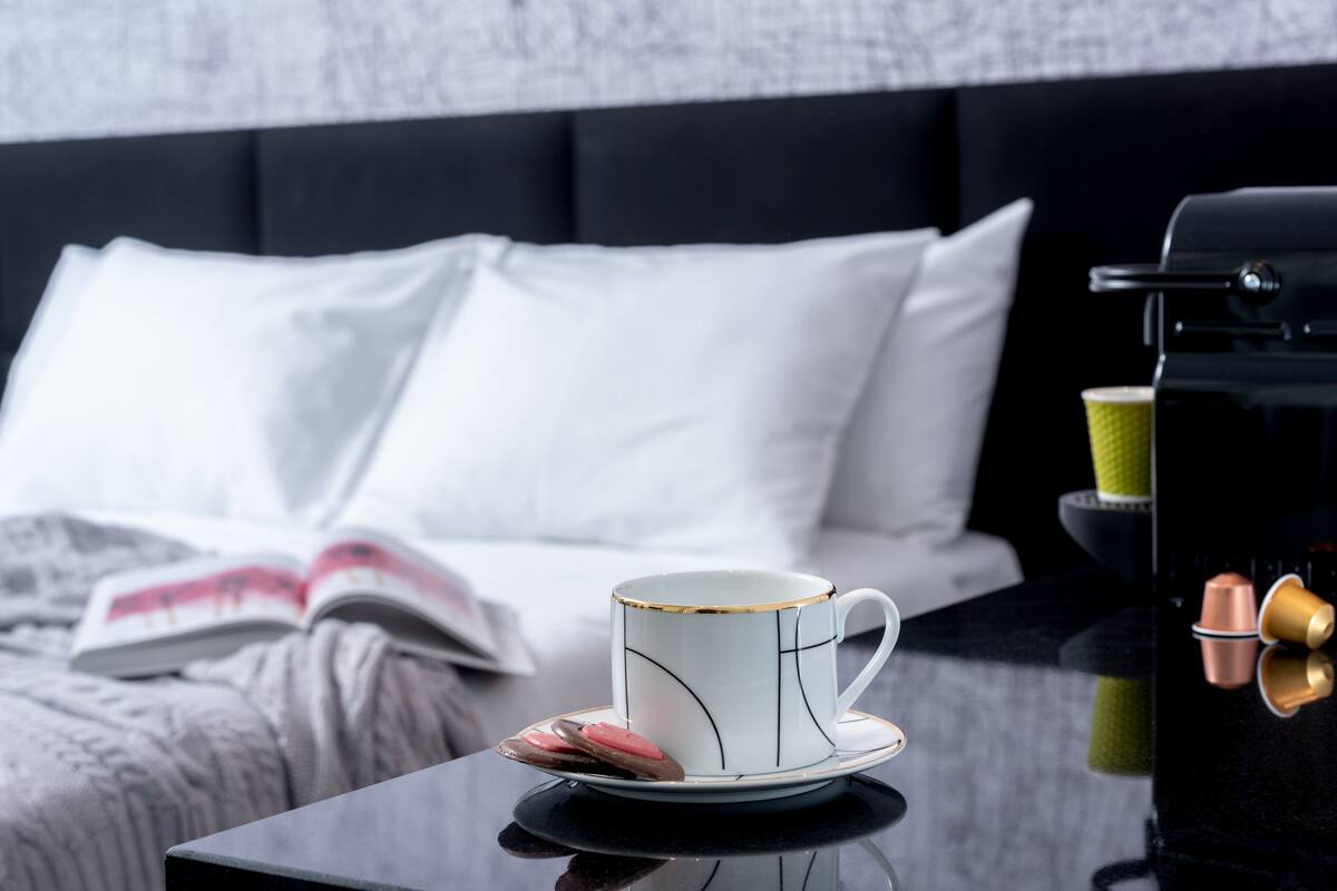 Ein ordentlich bezogenes Bett mit weißen Kissen und einem Buch darauf. Eine weiße Tasse mit goldener Verzierung auf einem Untertasse, und daneben eine Kaffeemaschine mit Tasse und Untertasse.