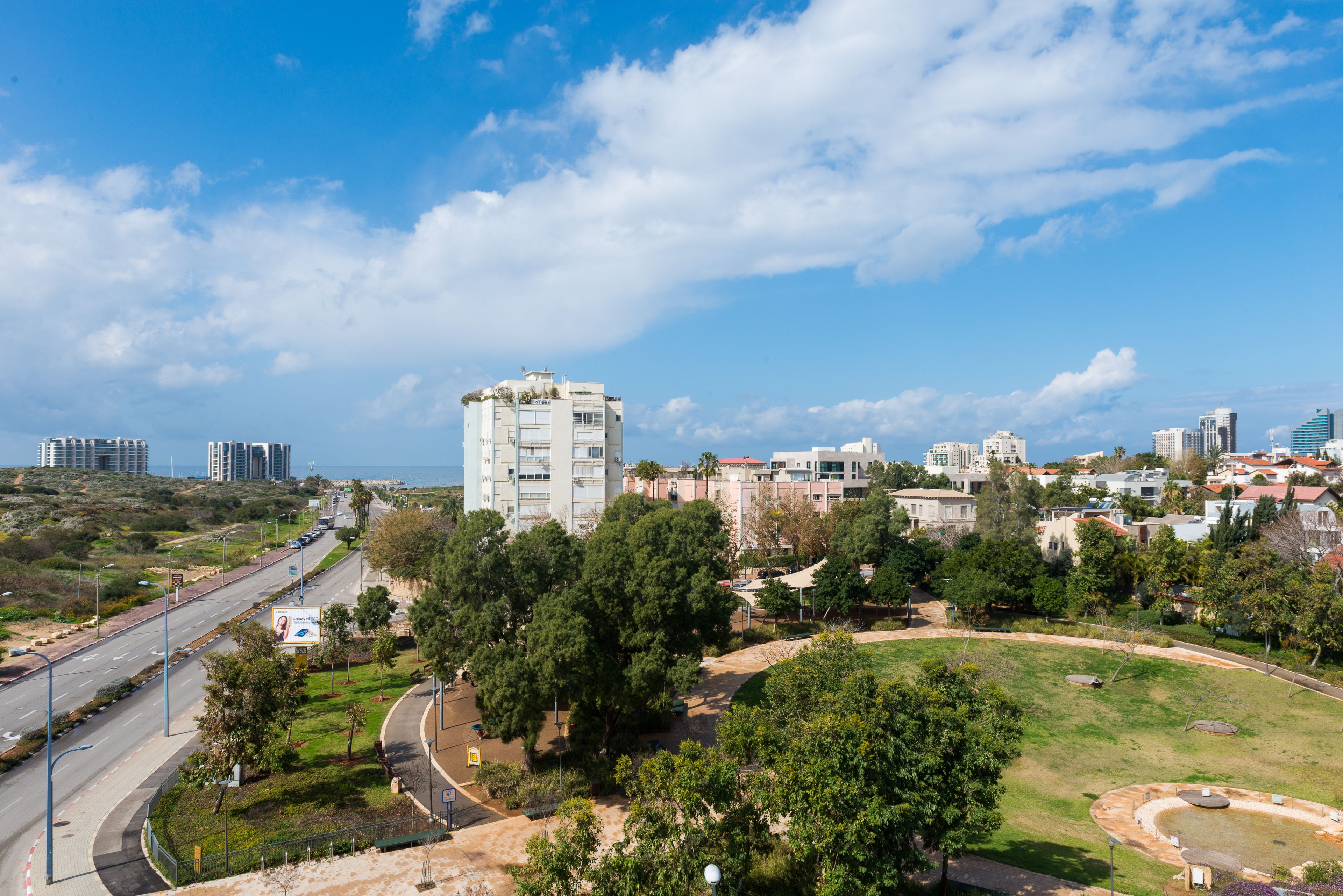 A cityscape with a prominent white building, a curved road, and a park area with trees and a small pond.