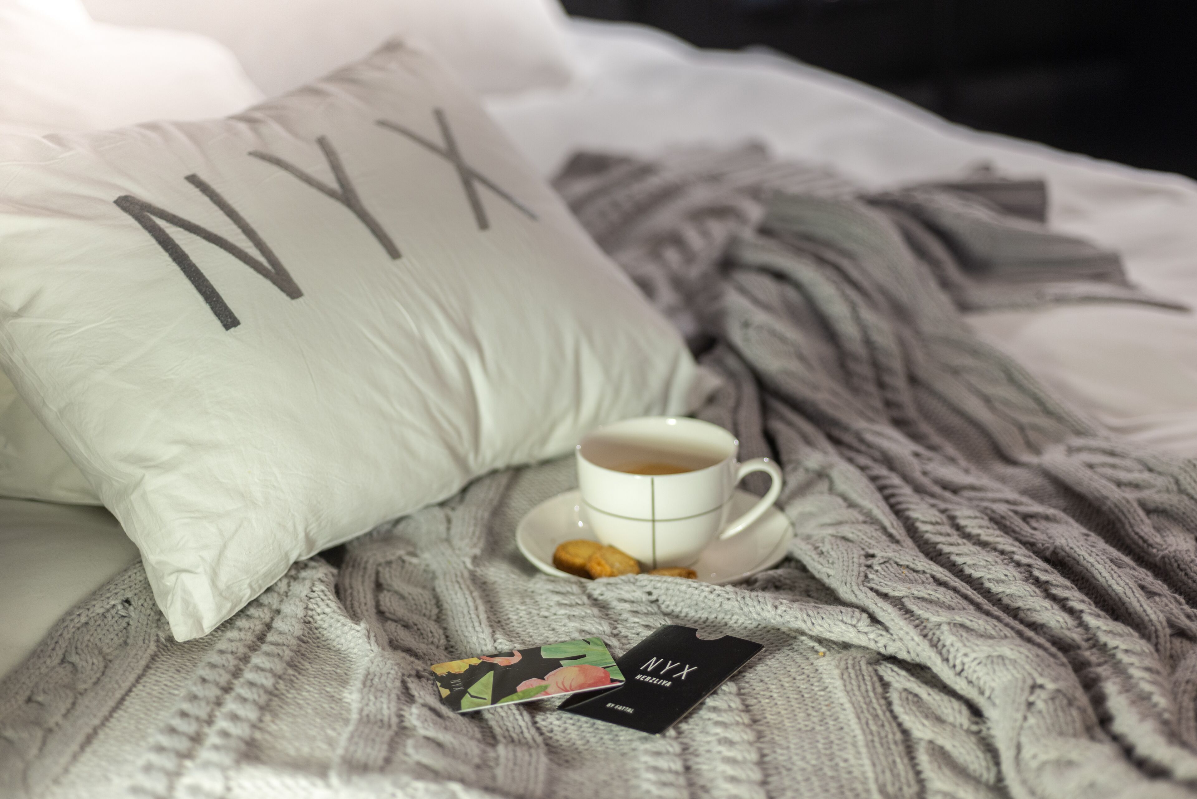 Una cama con un almohadón blanco, una taza de café, una tarjeta y una caja de bombones NYX.