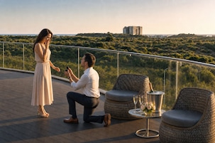 Un hombre le pide matrimonio a una mujer en un balcón con muebles de mimbre, una mesa con una botella y copas, y vistas a la ciudad.