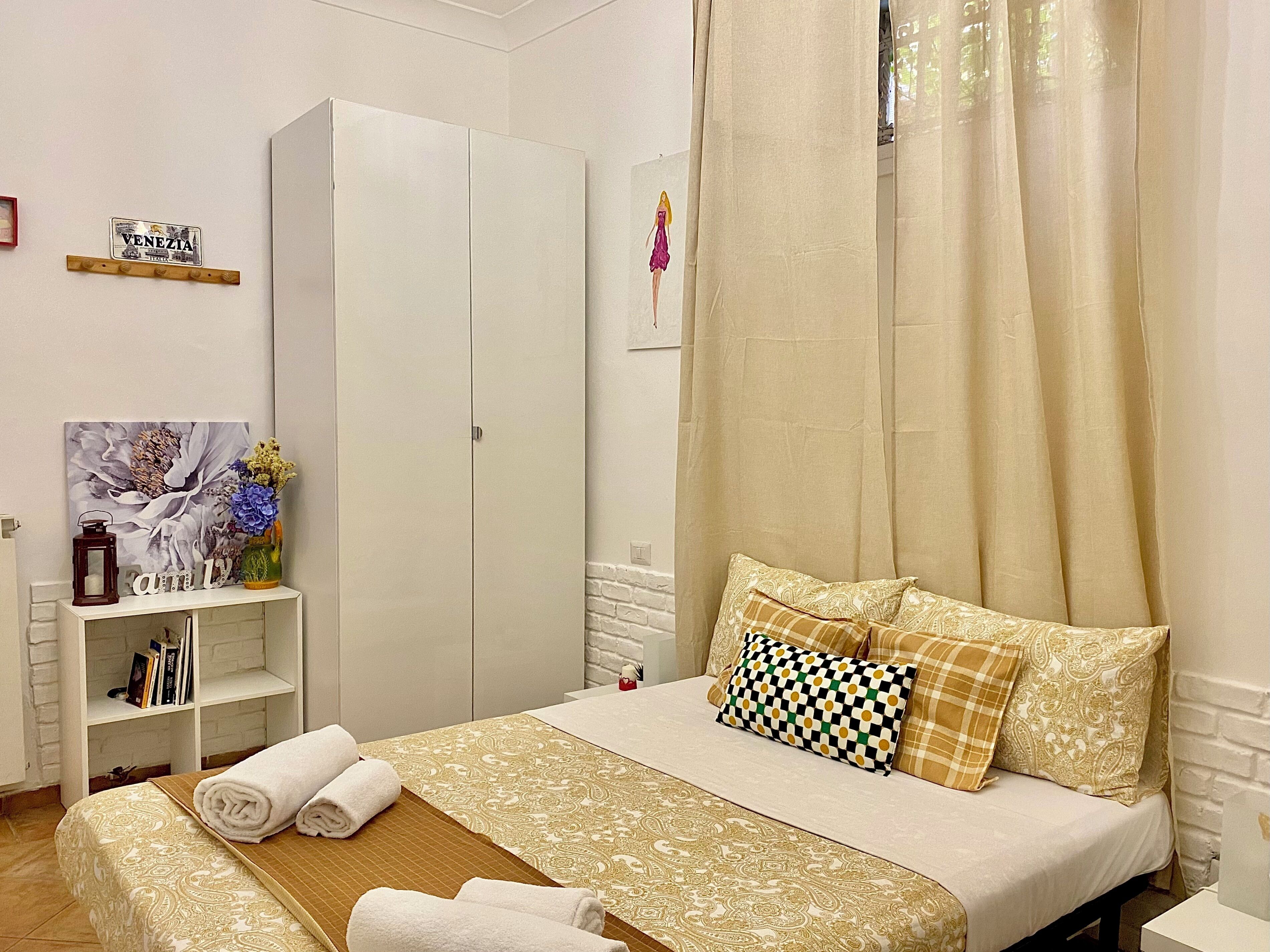 A neatly arranged bedroom with a bed, a wardrobe, a shelf with books, and a painting on the wall.