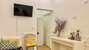 A living room with a flat-screen TV mounted on the wall, a sofa with patterned cushions, a yellow chair, a small table with a tray, and a decorative vase on a console table. - Domus Romana B & B (Rome)