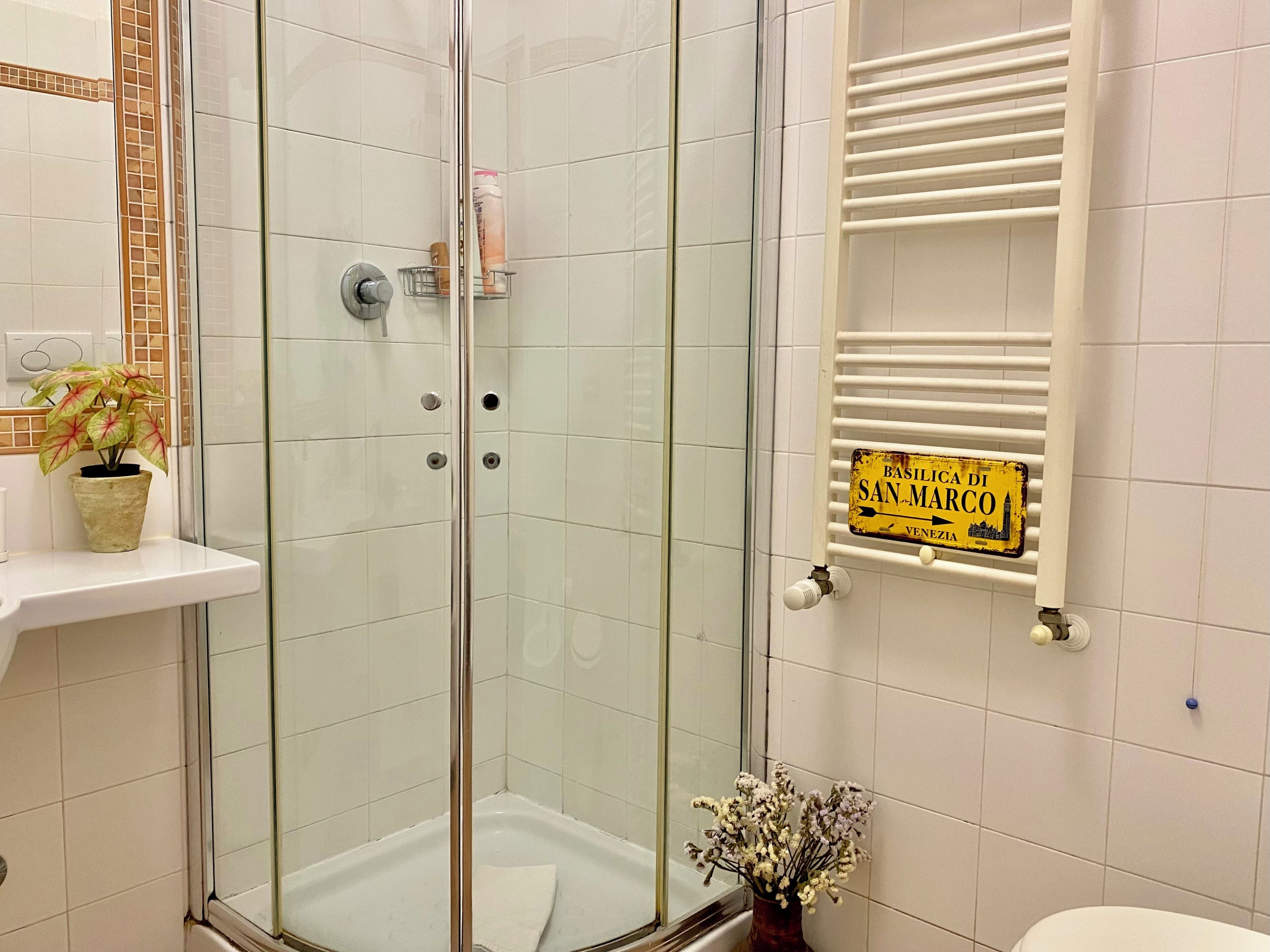 A bathroom with a glass-enclosed shower, a white sink with a potted plant, a towel rack, and a sign indicating the location as 'Basilica di San Marco, Venezia'.