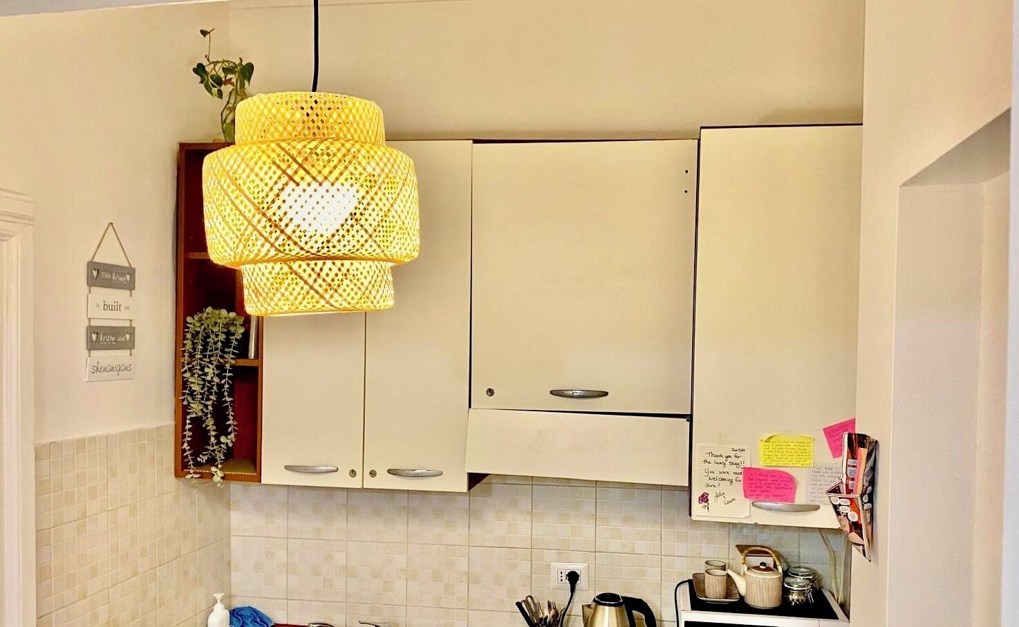 Una cocina con armarios color beige, una lámpara colgante amarilla y una pequeña estantería con utensilios de cocina.