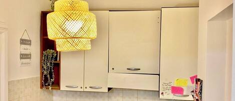 Una cocina con armarios color beige, una lámpara colgante amarilla y una pequeña estantería con utensilios de cocina.
