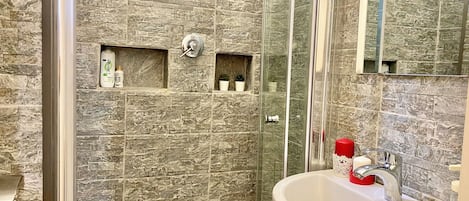 A bathroom with a stone-textured wall, a glass-enclosed shower, and a white sink with a red soap dispenser.