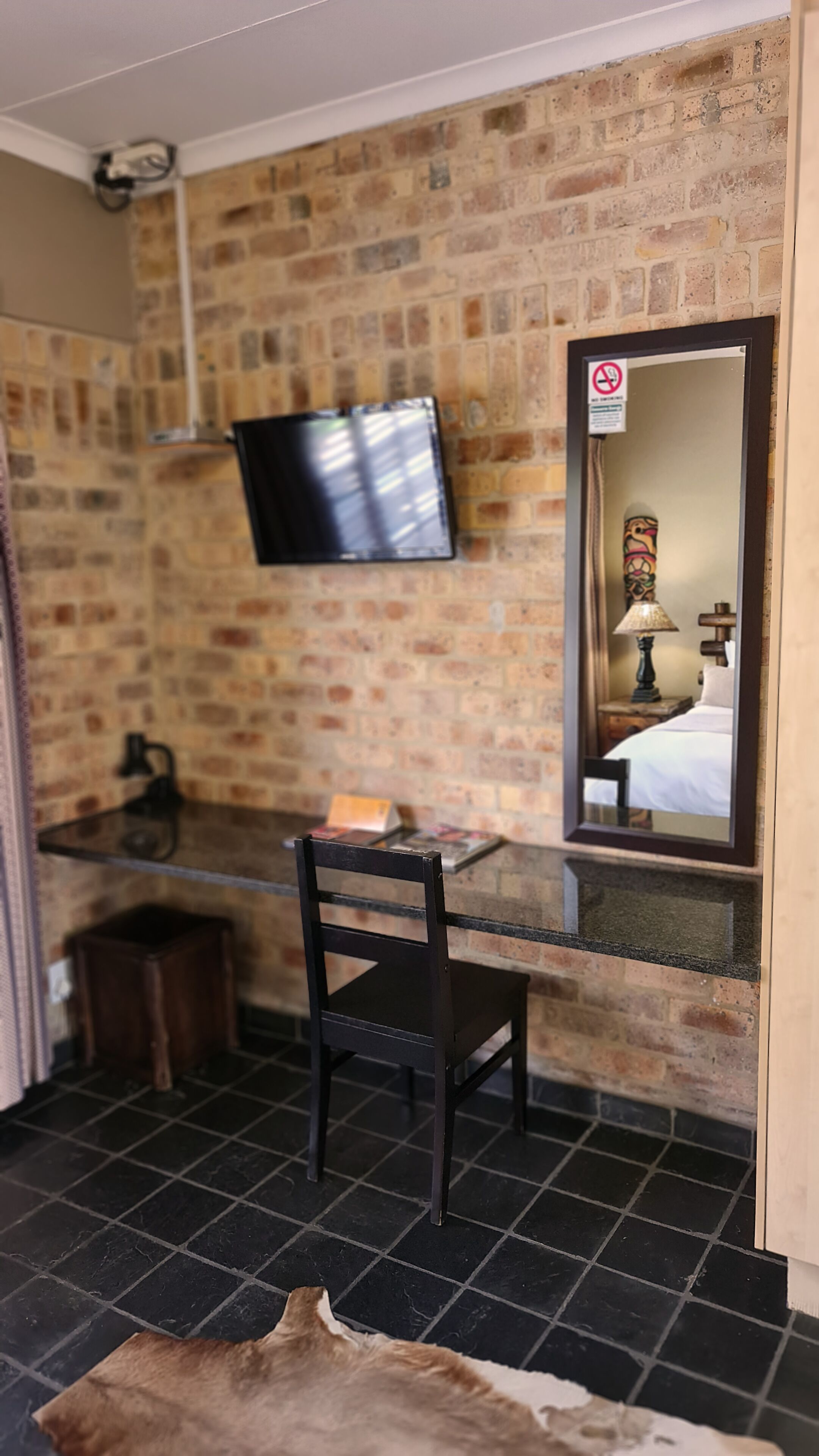 Une pièce avec un mur en briques, un téléviseur fixé au mur, un bureau avec une chaise, un petit tabouret et un miroir qui reflète un lit et une lampe.