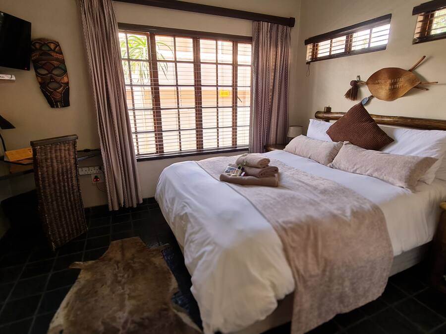A hotel room with a bed, a television, a window with curtains, a wall-mounted decorative item, and a small table with a lamp.