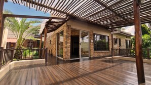 A covered outdoor area with a wooden deck, a brick building with glass doors, and a view of palm trees. - Klein Bosveld Guesthouse (Emalahleni)
