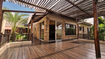 Área externa coberta com deck de madeira, um edifício de tijolos com portas de vidro e vista para palmeiras.
