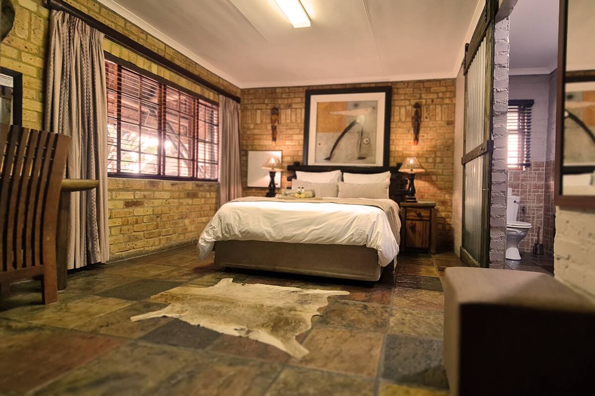 A bedroom with a large bed, a wooden bench, a rug, a brick wall, and a window with curtains.