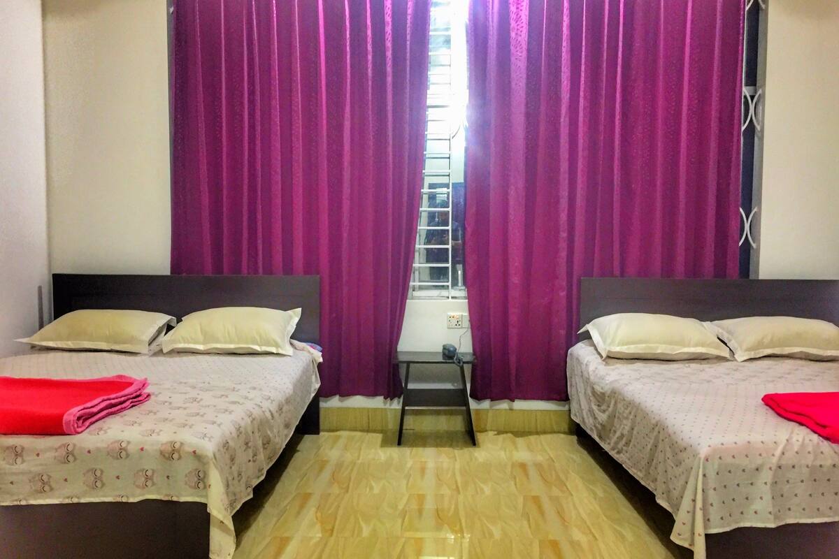 Une chambre avec deux lits, chacun étant équipé d’un oreiller et d’une couverture rouge. Il y a une fenêtre donnant sur un bâtiment à l’extérieur.