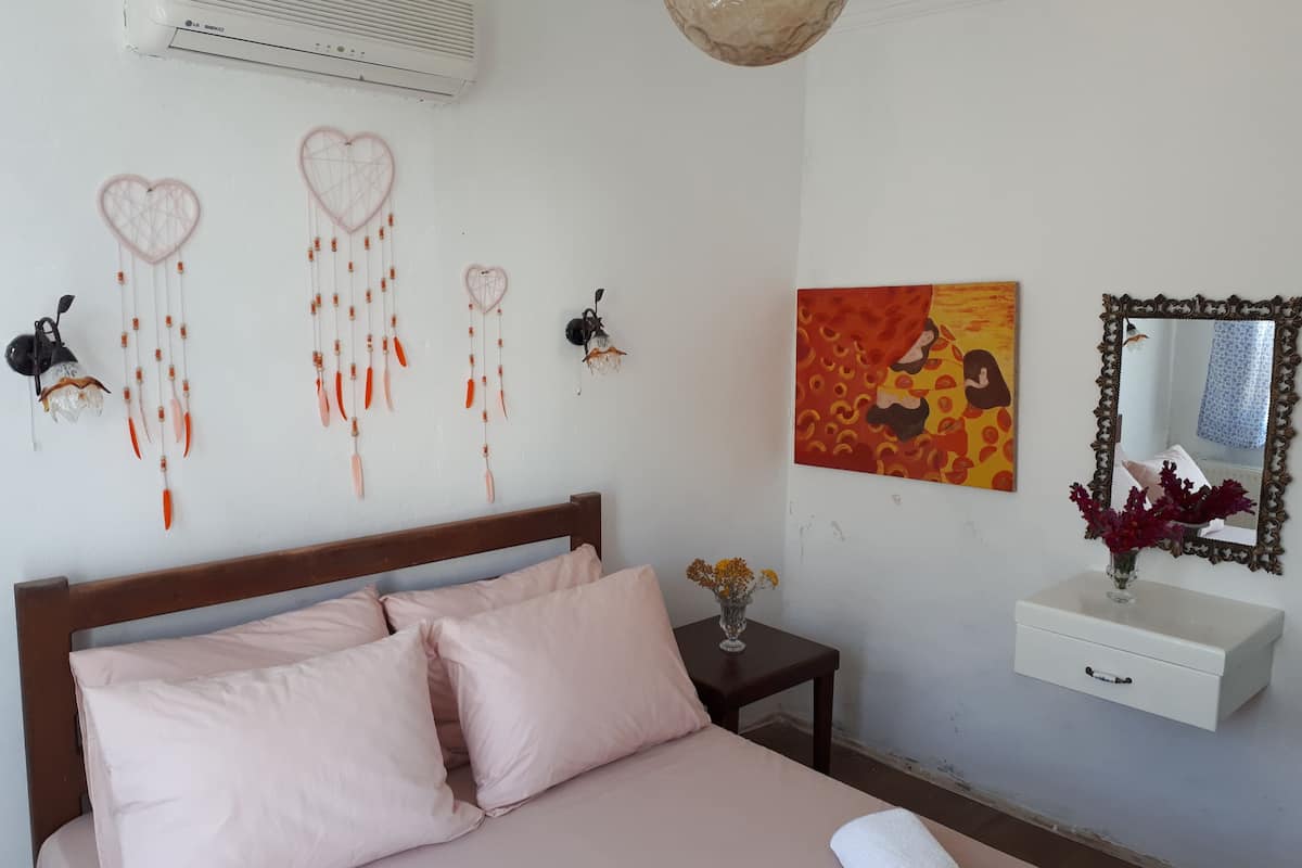 Un dormitorio con cama, mesita de noche, aire acondicionado instalado en la pared, adornos en la pared, un cuadro, un espejo y un florero con flores.