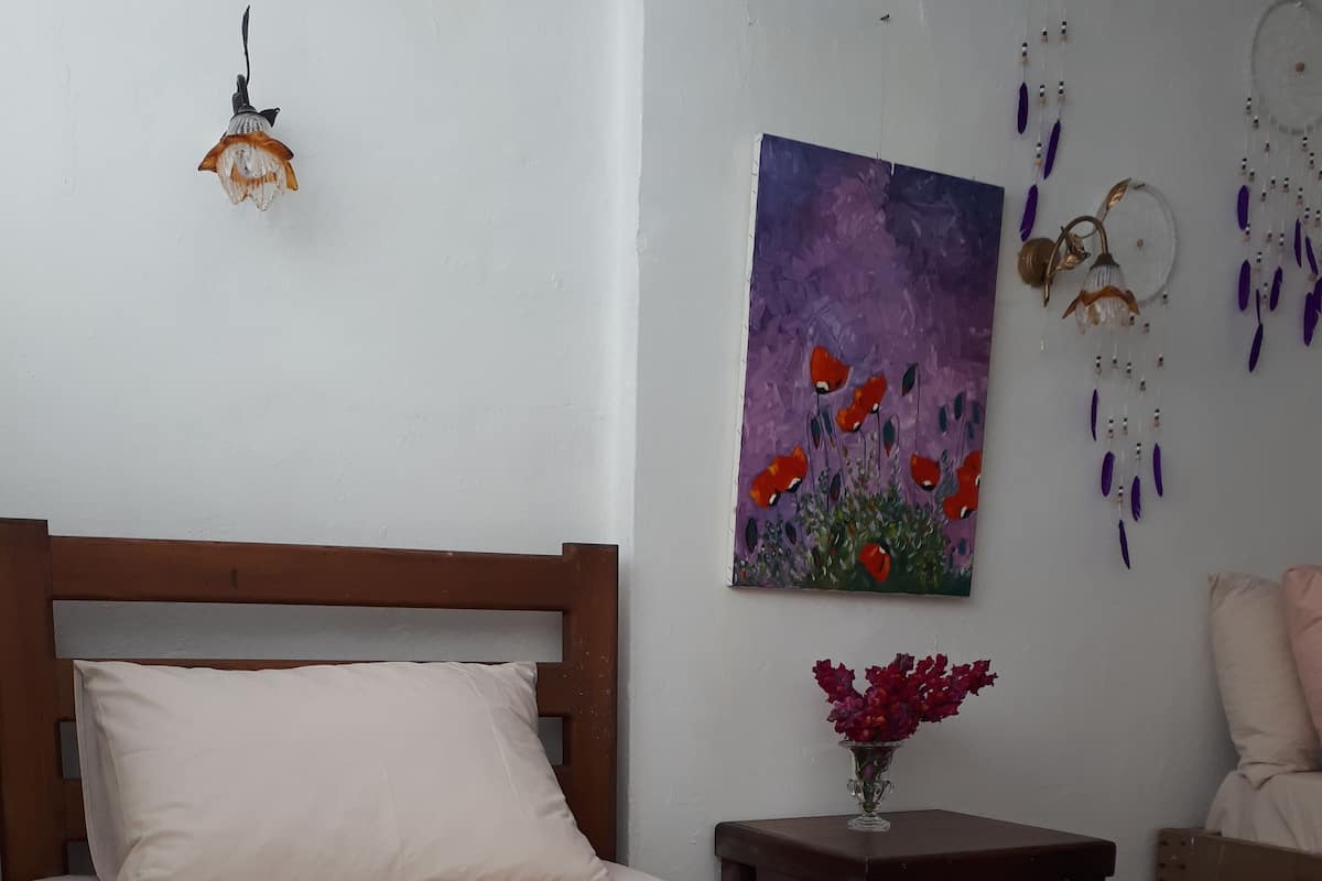 Una cama bien tendida con una colcha estampada con flores, una mesita de noche con un jarrón de flores, un cuadro en la pared y un atrapasueños.