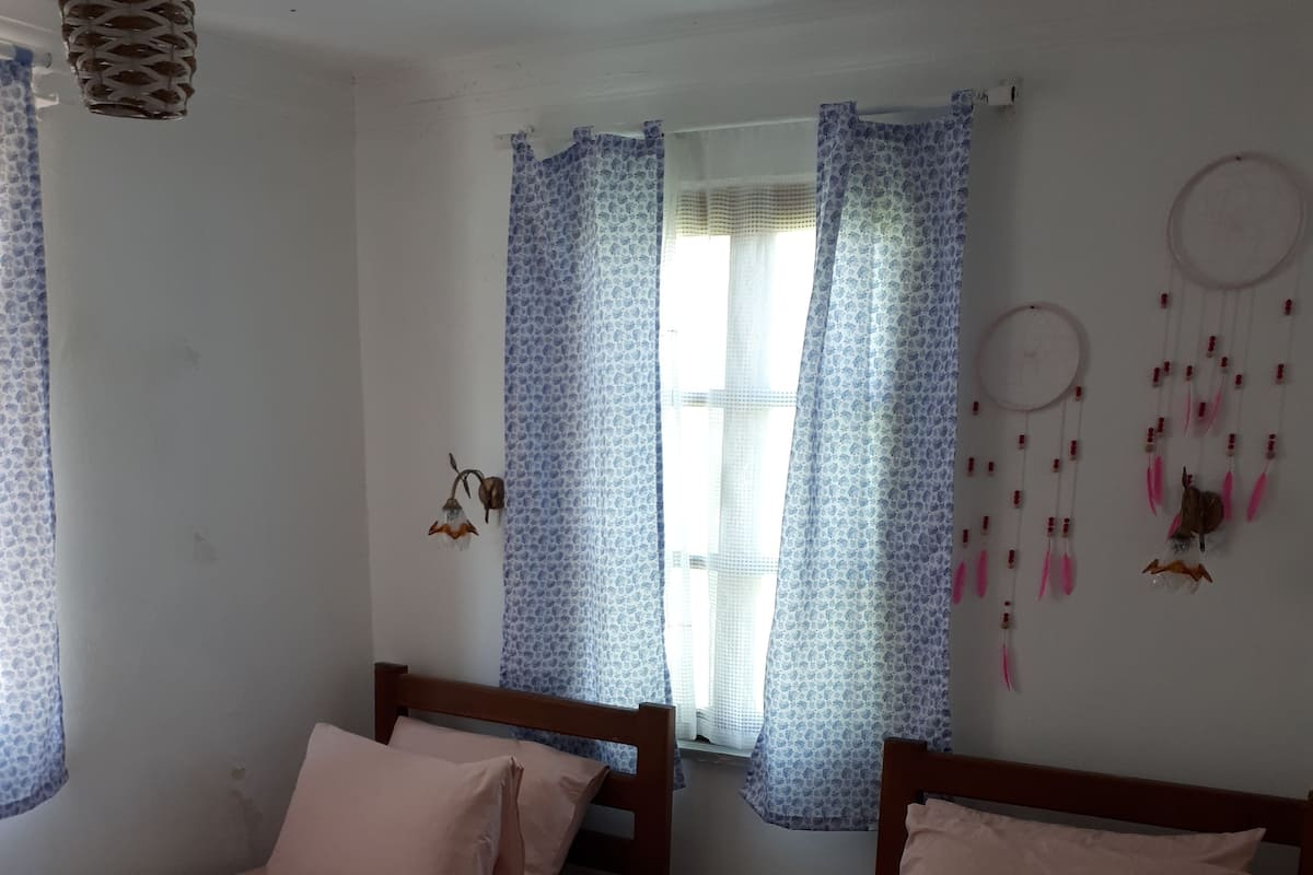 Una habitación con dos camas individuales, cada una con cabecero de madera y ropa de cama rosa. Hay una mesita con una toalla enrollada y un jarrón de flores. La habitación tiene cortinas con estampado y un atrapasueños en la pared.
