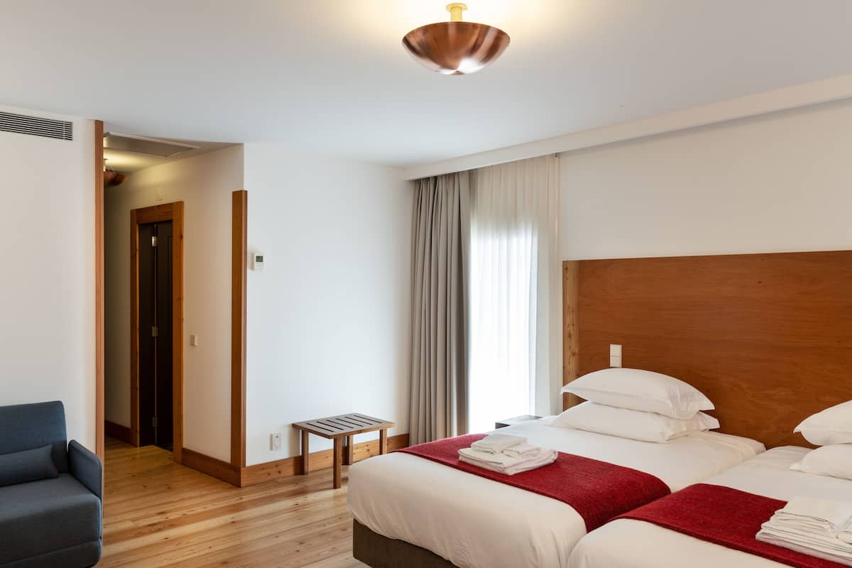 A hotel room with a wooden headboard, a bed with white linens, a red throw blanket, a small wooden side table, and a grey armchair.