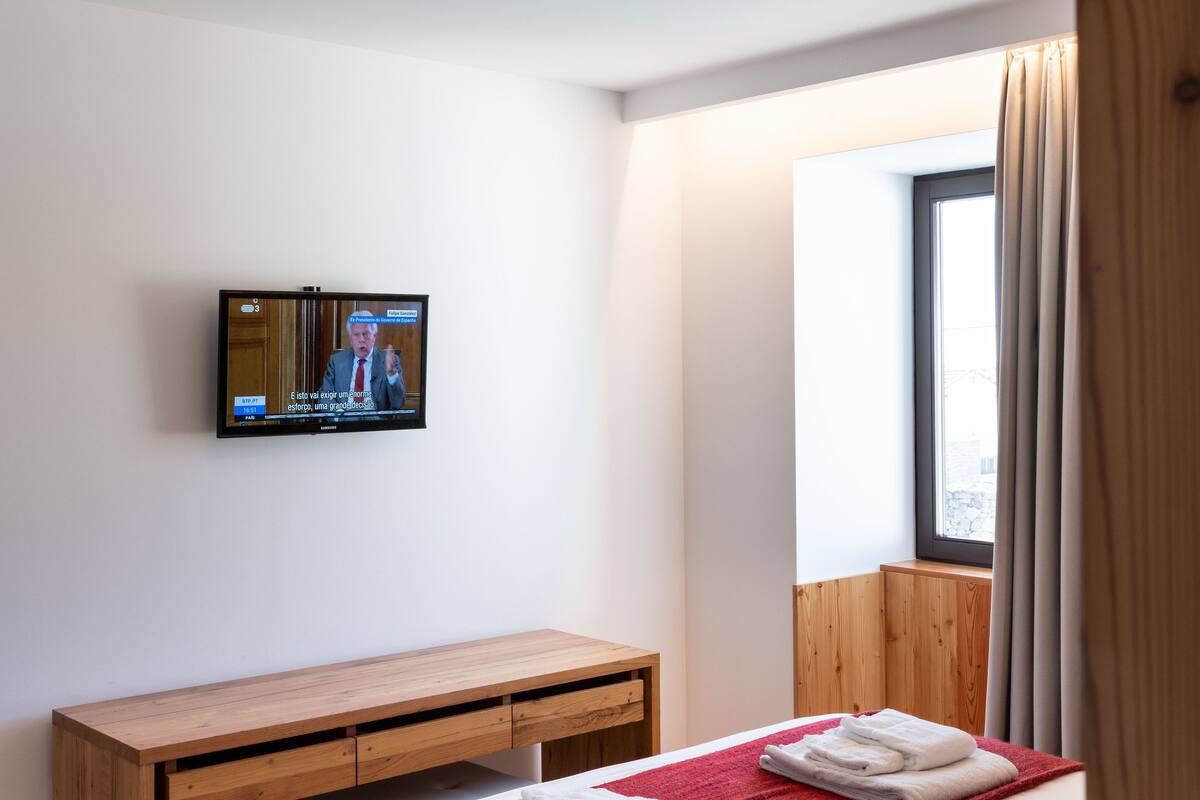 A hotel room with a bed, a wooden desk, a television mounted on the wall, and a window with curtains.