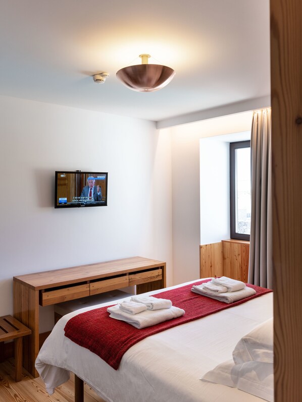 A hotel room with a bed, a wooden desk, a television mounted on the wall, and a window with curtains. - Casa de Santa Cruz (Torre de Moncorvo)