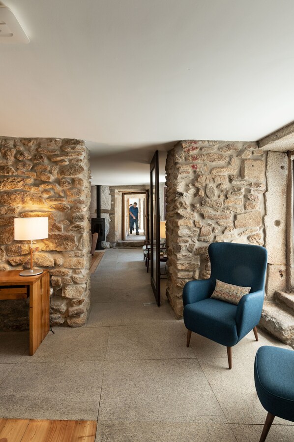 A stone-walled hallway with a blue armchair, a wooden side table with a lamp, and a fireplace in the background.