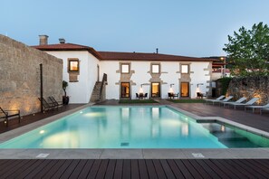A pool area with a stone wall, lounge chairs, and a building with multiple windows and doors.