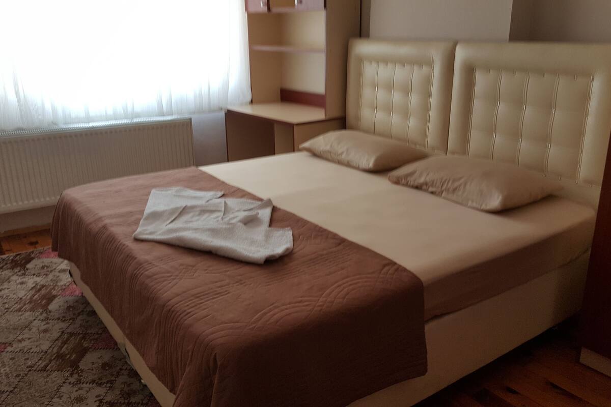 A neatly made bed with a brown bedspread and two pillows, a bedside table, a radiator, and a patterned rug.