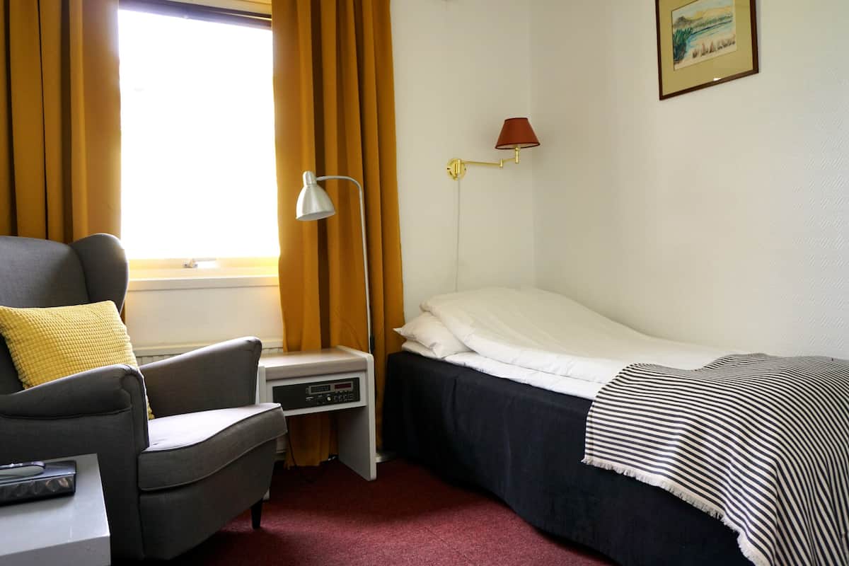 A hotel room with a single bed, a bedside table with a lamp, a chair with a yellow pillow, and a window with curtains.