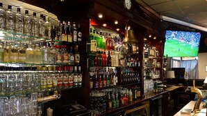 Um bar com prateleiras cheias de garrafas de bebidas e copos, uma televisão exibindo uma partida de futebol e um relógio na parede.