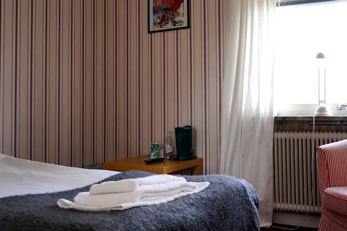 A hotel room with a bed, a striped armchair, a small table with a bottle and a cup, and a window with curtains.