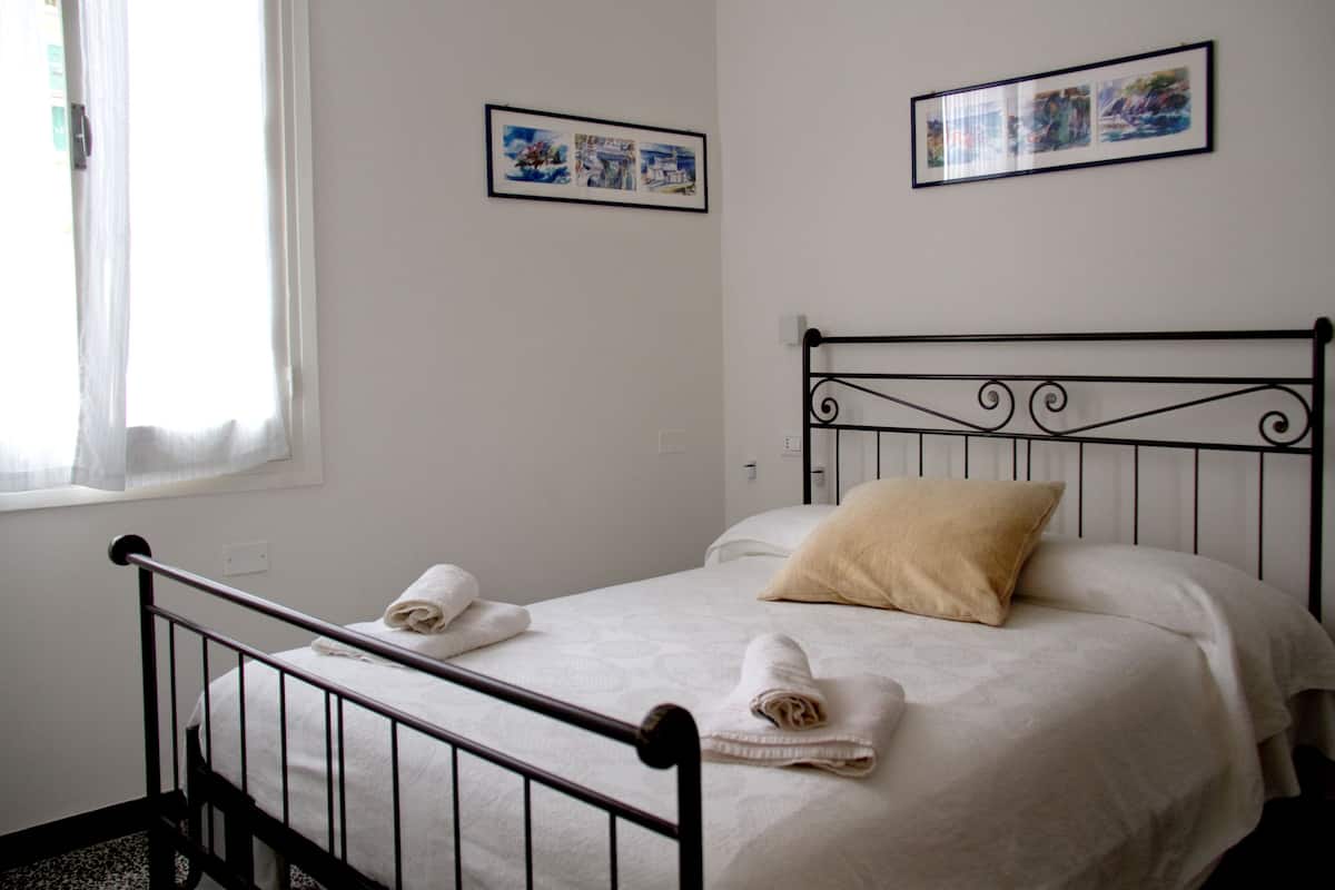 A neatly made bed with white linens and two rolled towels, a window with sheer curtains, and two framed pictures on the wall.