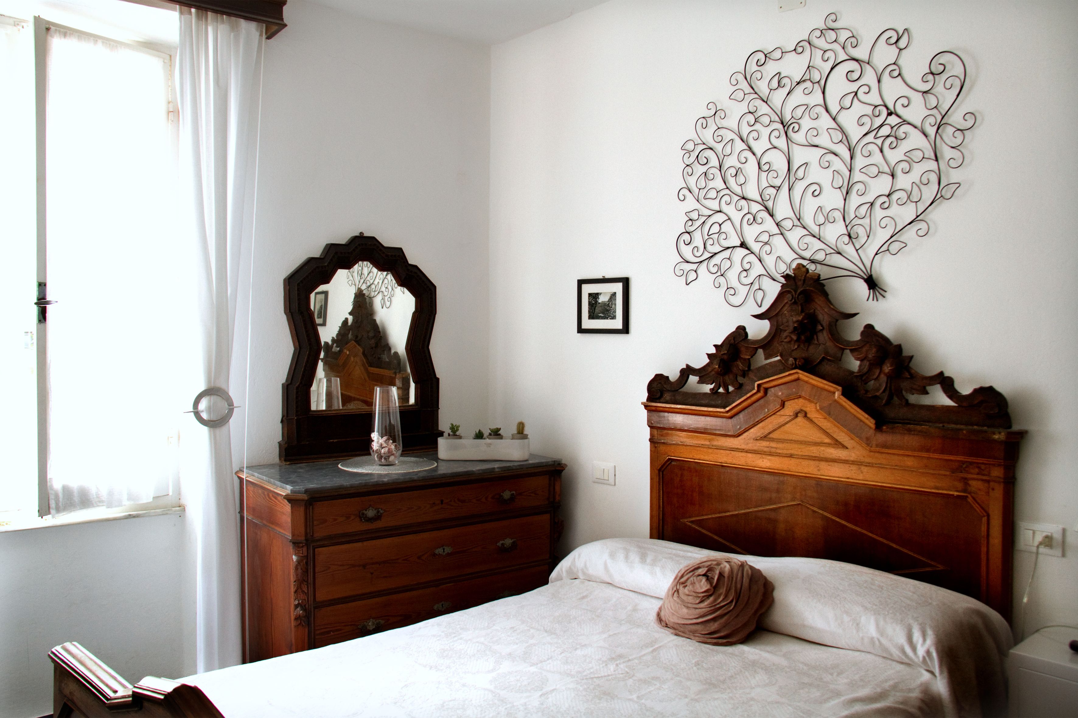 Une chambre à coucher avec une commode en bois, un miroir, un lit recouvert d’une courtepointe blanche et orné d’un oreiller marron, ainsi qu’un élément décoratif mural.