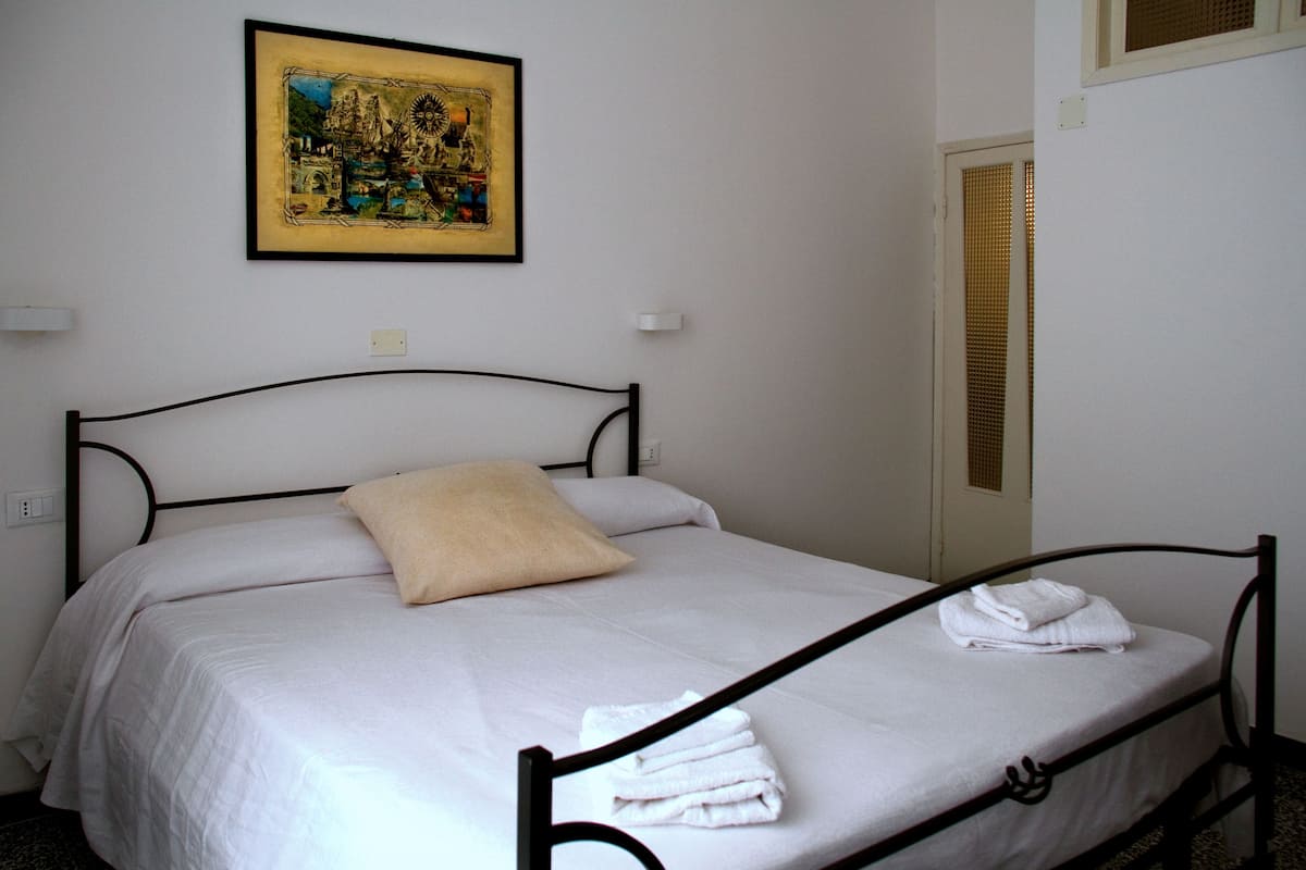 A single bed with white bedding and a beige pillow, a framed picture on the wall, and a window with blinds.
