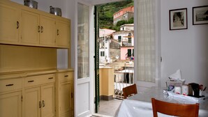 A room with a table, chairs, a yellow cabinet, and a view of buildings through an open door. - Guesthouse Rollando (Vernazza)