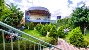 A building with a curved roof and a balcony, surrounded by greenery and a paved area with chairs. - Lebanon Hotel (Kigali)