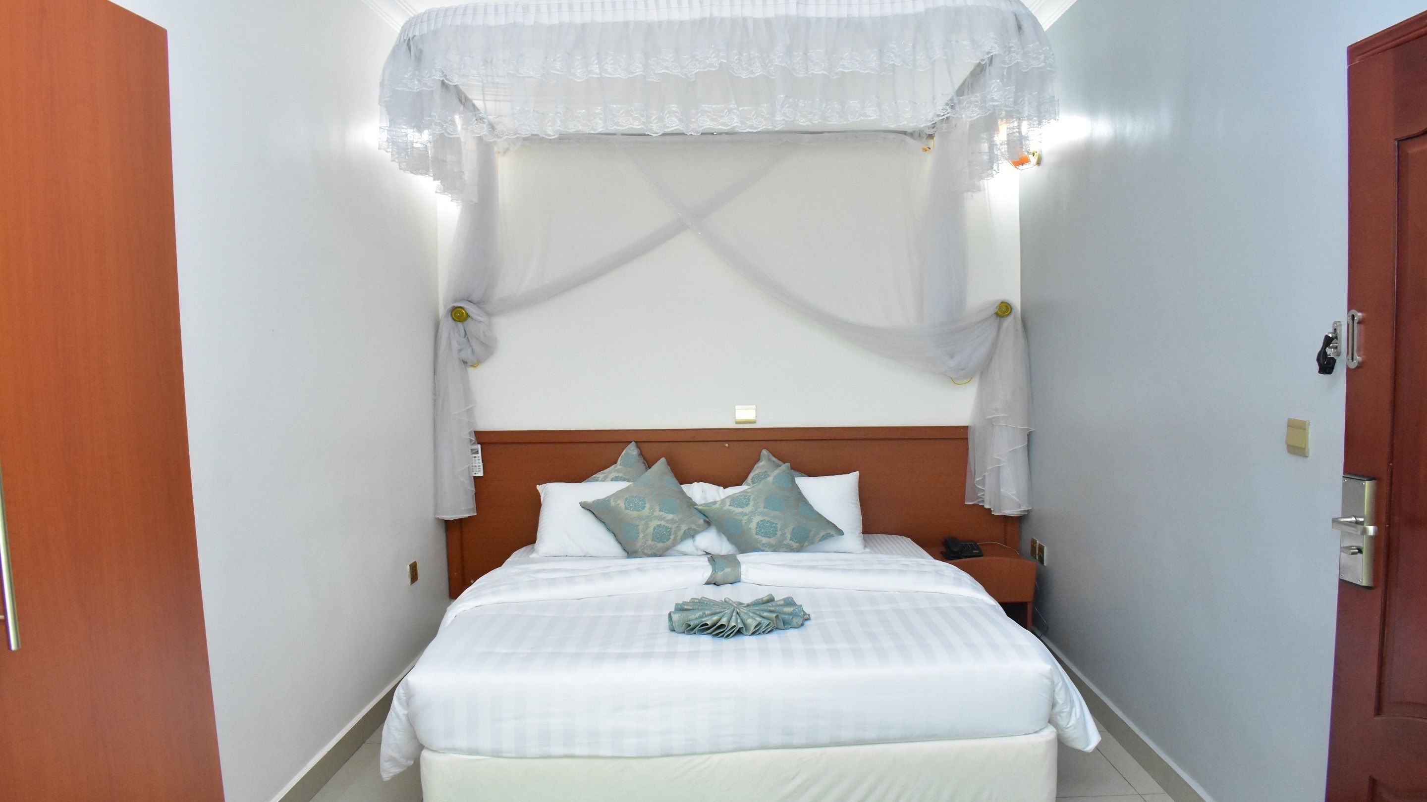 Una habitación de hotel con cama, cortinas blancas, cabecera de madera y mesita de noche.