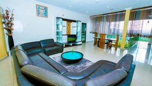 A modern living room with a sectional sofa, a coffee table, and a dining area with wooden chairs and a glass table. - Lebanon Hotel (Kigali)