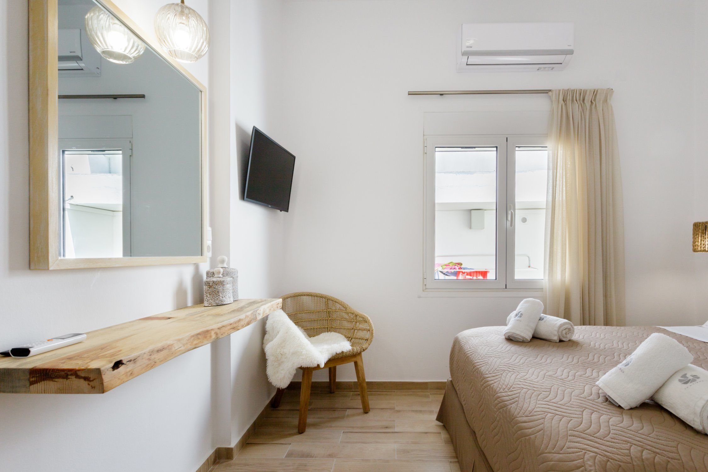 A hotel room with a wooden shelf, a wicker chair, a bed with a beige quilt, a flat-screen TV, and a window with curtains.
