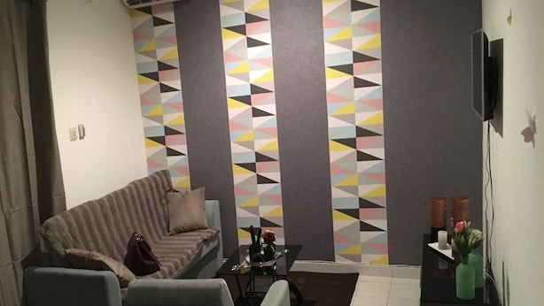 Een woonkamer met een bank, fauteuils, een salontafel en een televisie. Er is een behang met een patroon en een klein bijzettafeltje met kaarsen.