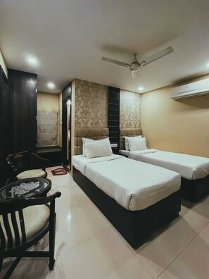 A hotel room with two single beds, a ceiling fan, a small table with chairs, and an air conditioning unit.