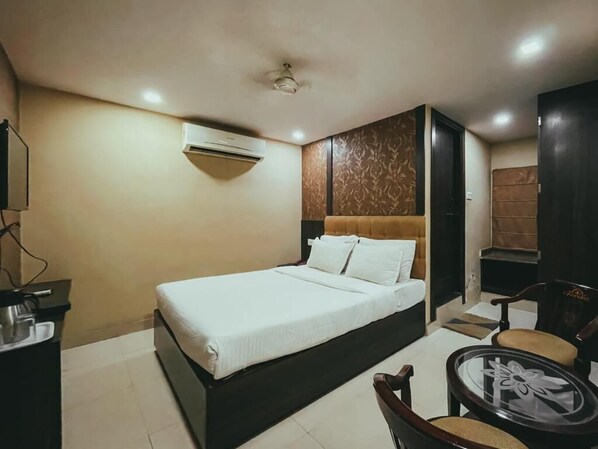 A hotel room with a bed, a desk with a chair, a small table, and an air conditioning unit.