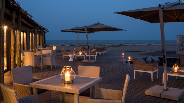A wooden deck with outdoor seating, tables, and umbrellas, overlooking the beach.