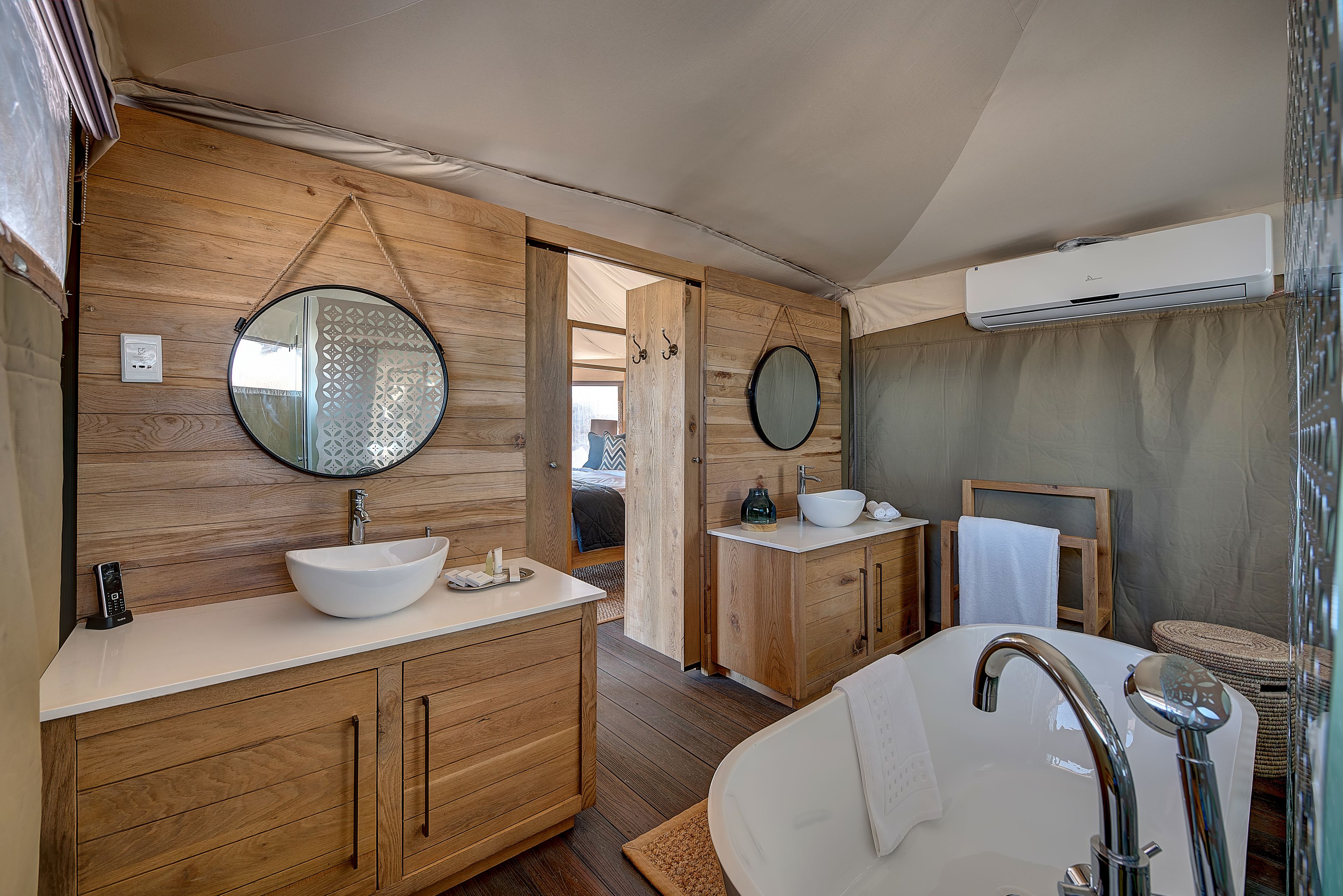A modern bathroom with wooden cabinetry, a large round mirror, a white sink, and a freestanding bathtub.
