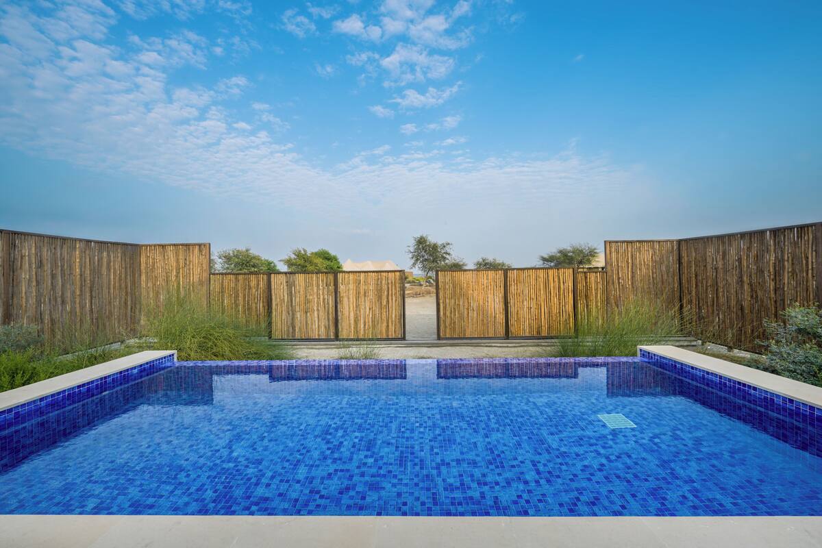 A rectangular swimming pool with blue mosaic tiles, surrounded by a wooden fence, and a clear sky above.