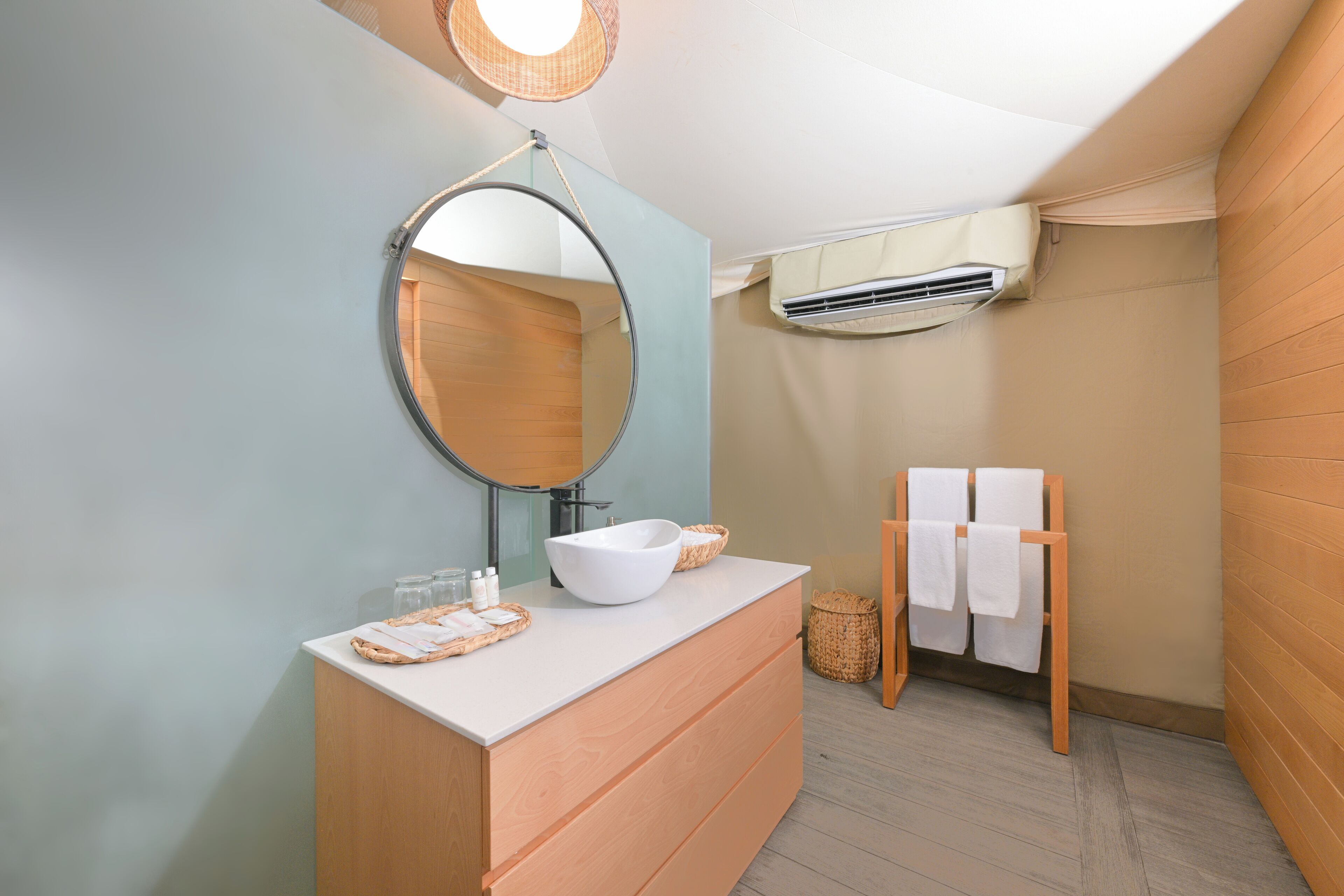 Un baño moderno con un espejo redondo, un lavabo blanco y un mueble de tocador de madera. Se aprecia un perchero con dos toallas y una cesta en el suelo.