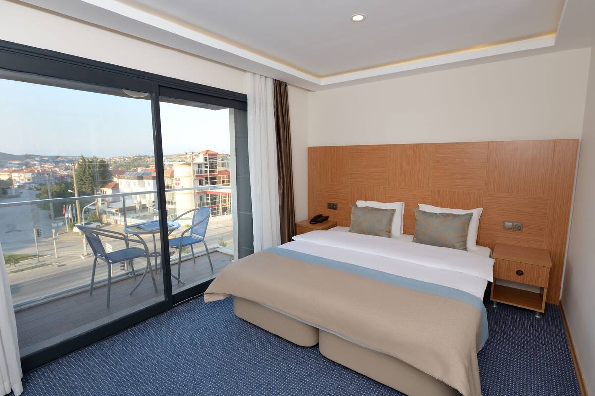 Habitación de hotel con una cama grande, cabecera de madera, balcón con sillas y vista a la ciudad.