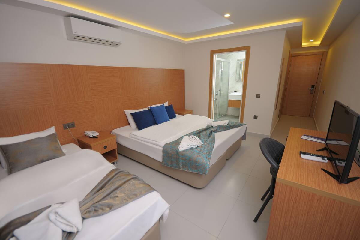 Habitación de hotel con dos camas, un escritorio con ordenador y un baño visible a través de una puerta de cristal.