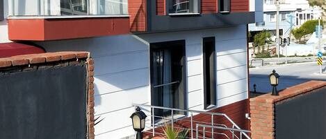 A modern multi-story building with a red and white facade, a balcony on the upper floor, and a paved entrance area.