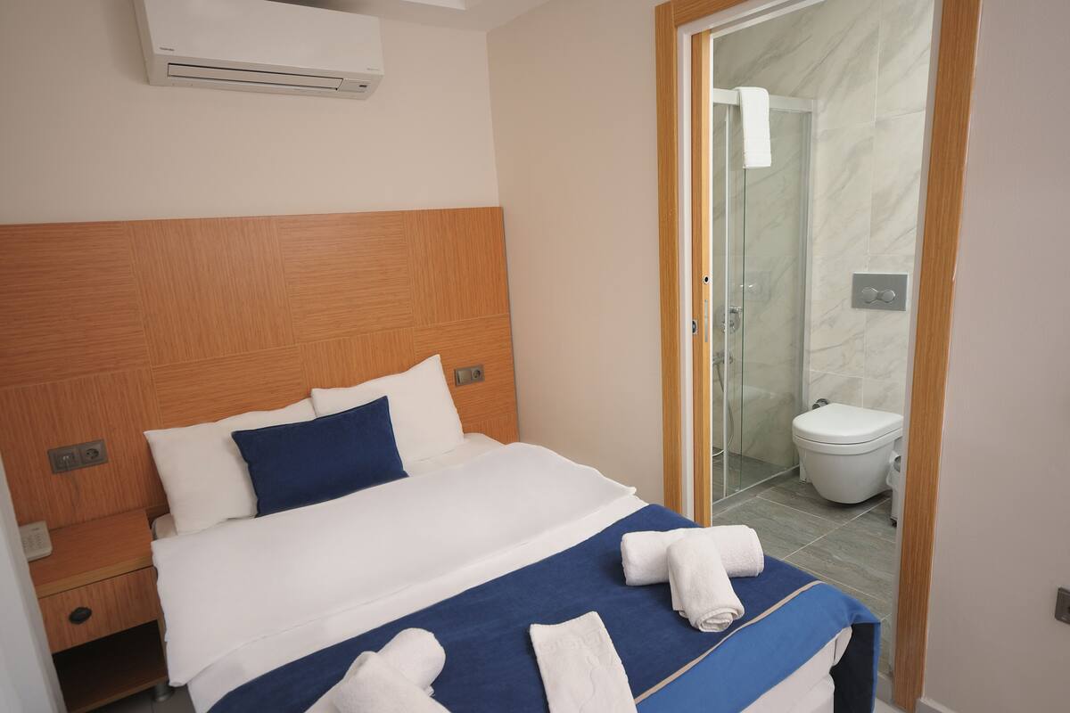 Habitación de hotel con cabecera de madera, cama grande con ropa de cama blanca y azul, baño con mampara de ducha de vidrio y un inodoro blanco.